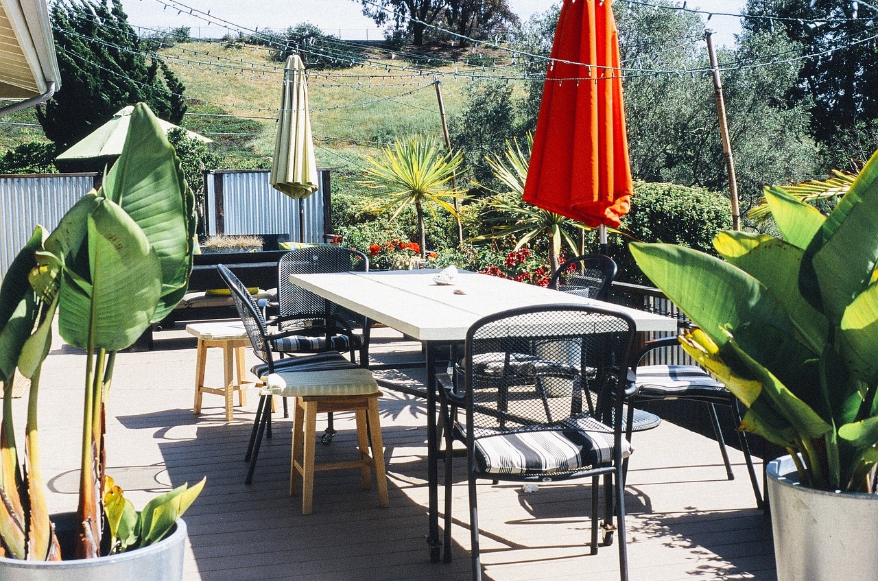 Table and chairs on an outdoor deck with umbrellas, elephant ear plants and overhead string lights